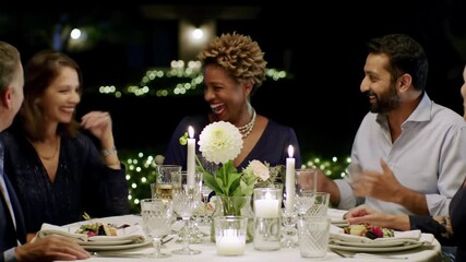 Diverse adults (30s-60s) laugh and converse at an outdoor night dinner, an exquisitely set table with dahlia, candles, and string lights, soft bokeh. Concept of shared joy