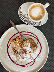 Cup of cappuccino served with slice of pumpkin strudel on ceramic plate with powdered sugar and berry syrup. Thanksgiving dessert break with warm coffee and cozy seasonal flavors