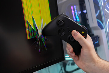 Aggressive gamer's hand holds a black controller, striking a computer monitor screen, causing severe cracks and colorful screen damage, depicting intense gaming frustration