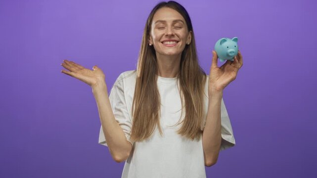 Young smiling woman holding blue piggybank and open palm gesture in purple studio; savings optimism.