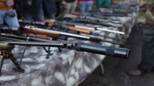 Confiscated rifles laid out on camo tables as people stand nearby observing seized weapons creating an atmosphere of arms trafficking investigation field inspection and evidence review