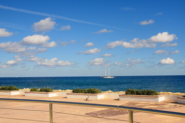 Blue summer sky with scattered white clouds over the Mediterranean Sea, featuring a sailboat and a concrete seaside promenade with potted green bushes.