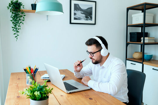 Young man wearing headphones sitting at desk working on laptop in modern office holding pen near mouth appearing focused during online meeting or remote work