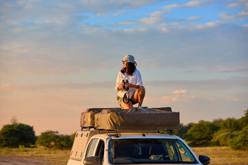 People on Safari theme: Solo Woman on the rooftop tent observing savanna for animals. at Golden Hour, Self-Drive Safari, Nxai Pan, Botswana.