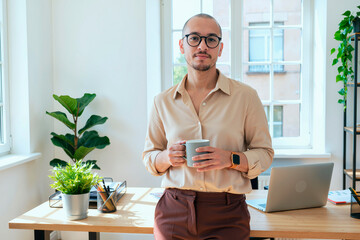 Portrait of young man standing in modern office holding coffee mug, wearing glasses, looking confidently at camera with laptop and potted plants on desk