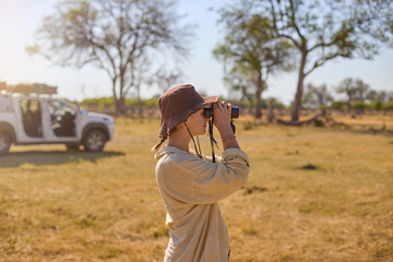Woman observes wildlife through binoculars beside an open 4x4 on a sunlit savanna. Ideal for: safari tourism, travel editorials, outdoor gear brands, and conservation storytelling.