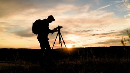 Male photographer with backpack uses camera tripod backdrop of stunningly beautiful sunset. Traveler with tripod camera takes beautiful landscape shots of stunningly beautiful sunny sky with clouds