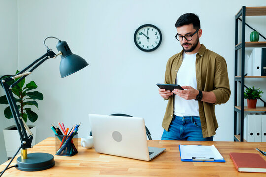 Young man standing at desk using digital tablet in modern office, laptop and documents on table, focused on screen, working on business project - Powered by Adobe