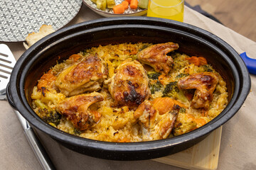 A cooked chicken and vegetable rice dish served in a round cast pot, placed on a wooden board with table elements visible around, showing a warm and hearty homemade meal in detail.