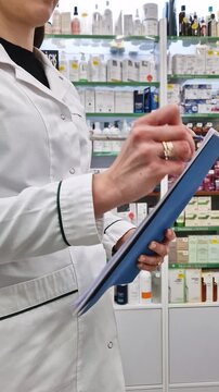 Female pharmacist or medical professional in a white lab coat holding a clipboard or folder and writing notes with a pen, like conducting inventory, checking stock, or fulfilling a prescription order