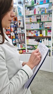 Female pharmacist or medical professional in a white lab coat holding a clipboard or folder and writing notes with a pen, like conducting inventory, checking stock, or fulfilling a prescription order