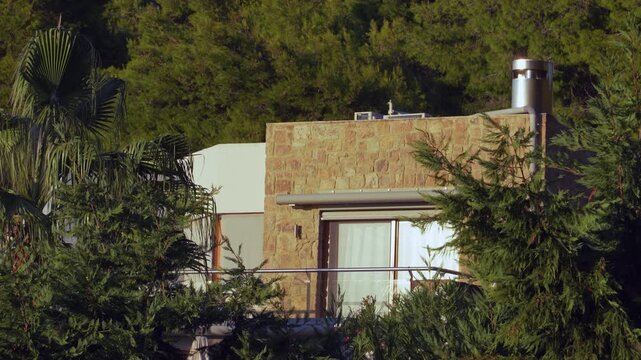 Modern stone clad villa with a glass balcony hidden amidst lush pine and palm trees in Greece, representing secluded eco friendly living and summer vacation