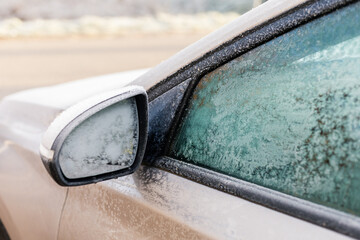 Frost coats the side mirror and window of a parked car in a cold winter landscape. The early...