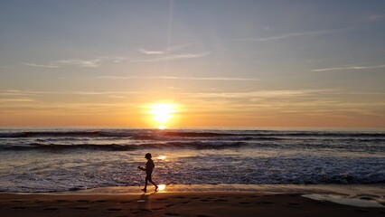 Silhouette of a child playing near ocean waves during sunset.