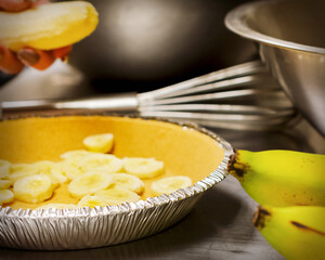 A chef's hand places sliced bananas into a graham cracker pie crust on a stainless steel counter, preparing a fresh banana cream pie in a local cafe.