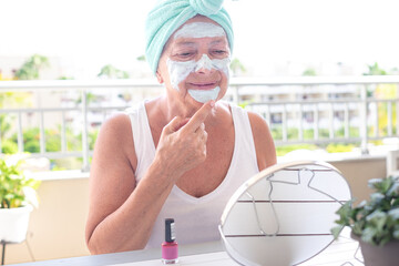 Portrait of a cheerful senior woman with a facial mask and a soft towel wrapped around her head looking at herself in the mirror, DIY, positivity, self-care concept