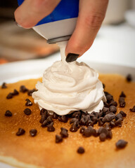 A chef adds a swirl of whipped cream from a can onto a fluffy pancake sprinkled with mini chocolate chips, creating a sweet breakfast.