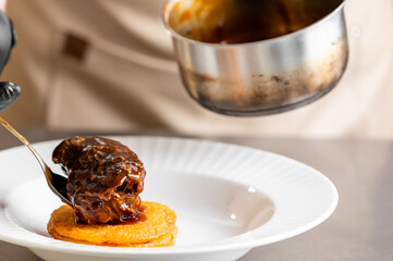 A close-up shot of a chef plating a serving of rich, slow-braised beef cheeks in a dark, glossy sauce on top of a round, golden base in a white bowl.