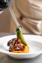 A chef carefully places a whole green jalape&ntilde;o pepper onto a gourmet braised beef dish with tweezers, highlighting culinary precision and modern food styling