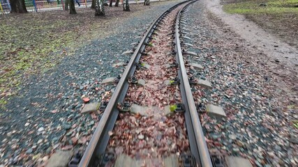 closeup narrow gauge rails curving through park leaf-littered sleepers and gravel under overcast sky, handheld follow shot evokes urban photographer wandering, subtle rust texture and tranquil mood - Powered by Adobe