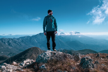 Obraz premium A lone hiker sits on a rocky ridge, quietly taking in the vast mountain landscape beneath a clear, teal-tinted sky.