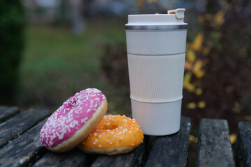 Two donuts, one pink and one orange, sit next to a warm drink cup on a weathered wooden table in a...