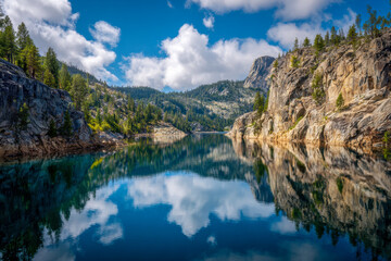 Fototapeta premium Tranquil mountain lake surrounded by rugged rocky cliffs and dense pine forests reflecting fluffy white clouds in clear blue water under a bright sky