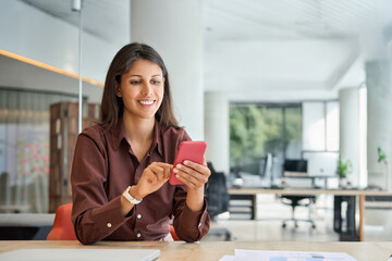 Hispanic business woman manager using cell phone mobile app, laptop. Smiling Latin or Indian student girl holding smartphone sitting in office or modern library, working online on gadget, copy space.