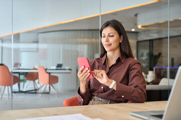 Young Indian business woman manager ceo using cell phone mobile app laptop. Smiling Latin Hispanic mature female businesswoman holding smartphone in office trading, reading, working online. Copy space