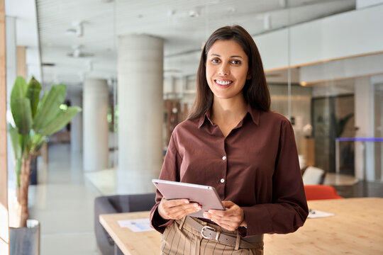 European businesswoman CEO using fintech tab application standing at workplace in office. Latin Hispanic mature professional business woman holding digital tablet for trading, looking at camera