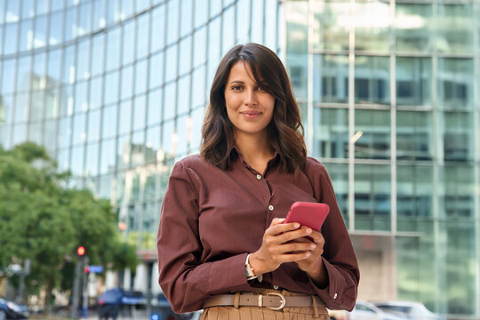 Portrait young latin hispanic business woman using smartphone cellphone financial trading app. Indian female entrepreneur businesswoman holding mobile cell phone for work outdoors looking at camera