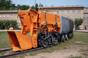An antique, bright orange motorized mine car with a large hydraulic shovel (loader) attached to the front, followed by a gray tipping wagon, displayed outdoors.