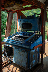 A wide shot of the abandoned, heavily rusted industrial control cabin used for mining machinery, showing the blue console and red structural framework in the sunlight.