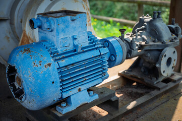 A heavy-duty blue electric motor coupled to a gray centrifugal pump or turbine, showing clear signs of weathering, rust, and industrial wear in an outdoor setting.