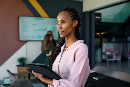 Thoughtful black young businesswoman with tablet in office with colleagues in background