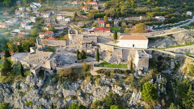 Aerial view of historic Klis Fortress in Croatia, bathed in warm golden hour light, overlooking the surrounding town and rugged landscape.