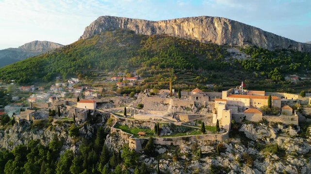 Aerial view of the historic Klis Fortress in Croatia, bathed in warm golden hour light, nestled beneath rugged mountains. A timeless blend of nature and heritage.