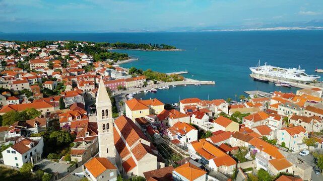 Aerial view of Supetar, Brac Island, Croatia. Charming red-roofed houses line the Adriatic coast, with a ferry docked in the bustling port under a clear sky.