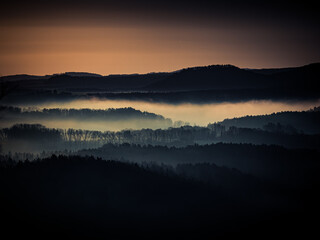 Moody sunrise over layered hills covered in dense morning fog. Soft blue shadows contrast with warm orange tones, creating a peaceful and atmospheric landscape with strong depth and texture.