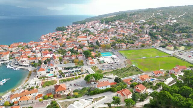 An aerial view of Postira, Brac Island, showcasing its charming red-roofed houses, turquoise Adriatic waters, and surrounding lush hills under a cloudy sky.
