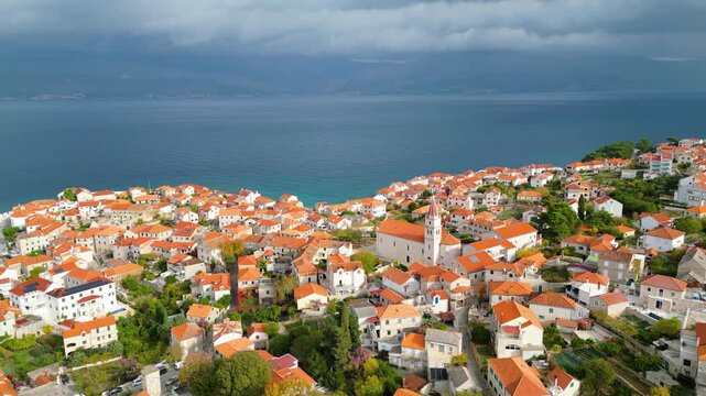 Aerial view of Postira, Brac Island, Croatia. Terracotta roofs meet the deep blue Adriatic under dramatic skies. A picturesque harbor town, blending history with modern tourism.