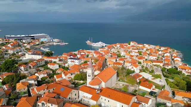 Aerial view of Postira, Brac Island, Croatia. Terracotta roofs meet the deep blue Adriatic under dramatic skies. A picturesque harbor town, blending history with modern tourism.