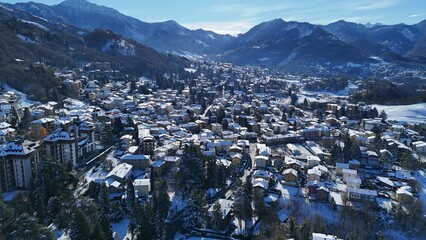A beautiful aerial drone shot of Barzio blanketed in fresh winter snow, located in the Valsassina valley of Lombardy, northern Italy. The image highlights alpine homes, snowy landscapes, mountain ridg
