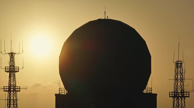 Silhouetted doppler radar and communication antennas rising against a vibrant sunset, highlighting the contrast between technology and nature
