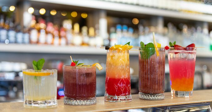 Five vibrant, colorful cocktails lined up on a sleek bar counter, each garnished with fresh fruit, mint, and citrus twists. ty and craftsmanship against a softly blurred backgound - Powered by Adobe