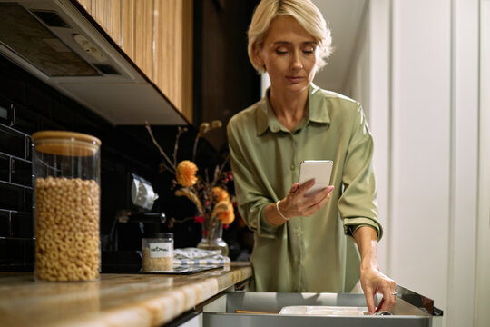 Middle aged Caucasian woman standing in modern kitchen holding smartphone in one hand while opening drawer with other hand, focusing on multitasking during daily routine