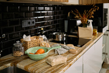 Kitchen countertop displaying assortment of groceries including oranges, bread, pasta, glass jar with nuts, woven basket, colander, and paper bag with dried flowers in background