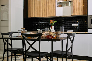 Modern kitchen interior featuring black dining table with empty chairs, countertop displaying various glass jars with food, coffee machine and vase with flowers on background