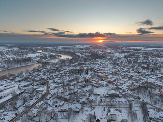 Aerial winter sunset view of small European town with snowy rooftops, winding river and dramatic colorful sky, perfect for travel, tourism or city background