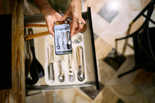 Caucasian young adult woman holding smartphone above open kitchen drawer, photographing organized cutlery tray with various utensils, hands and phone visible from overhead perspective - Powered by Adobe